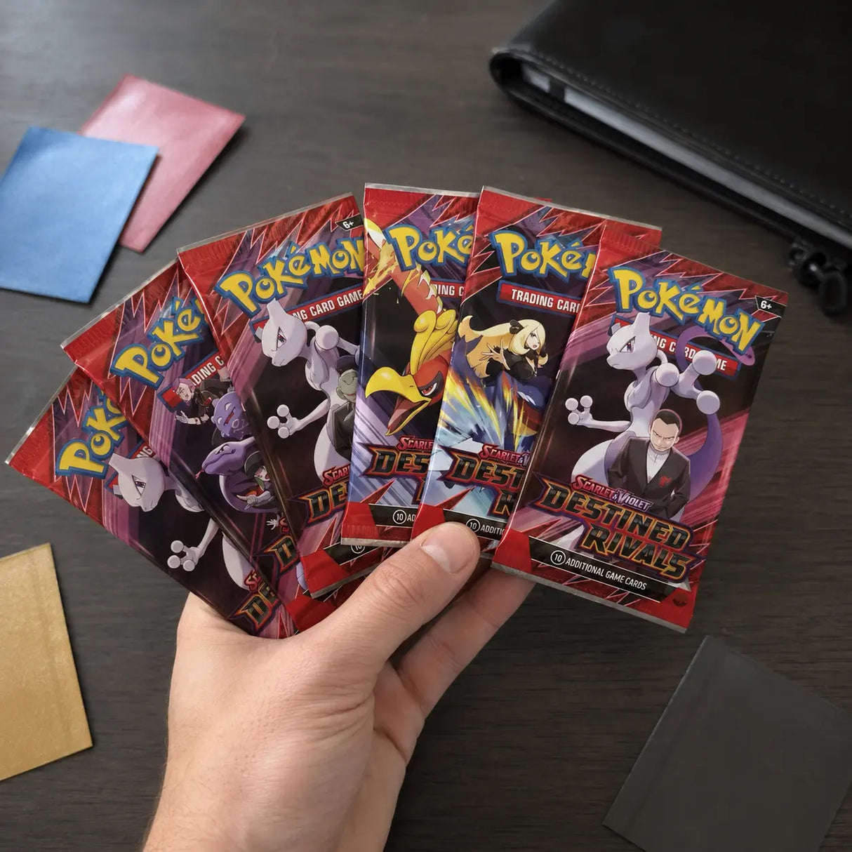 Hand hält 6 Destined Rivals Booster Packs aufgefächert – alle verschiedenen Artworks