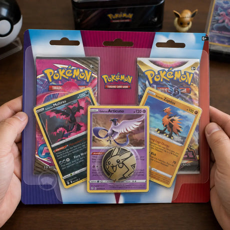 Hände halten Pokemon Galarian Birds Trio 2-Pack Blister auf Holztisch mit Evoli-Figur im Hintergrund