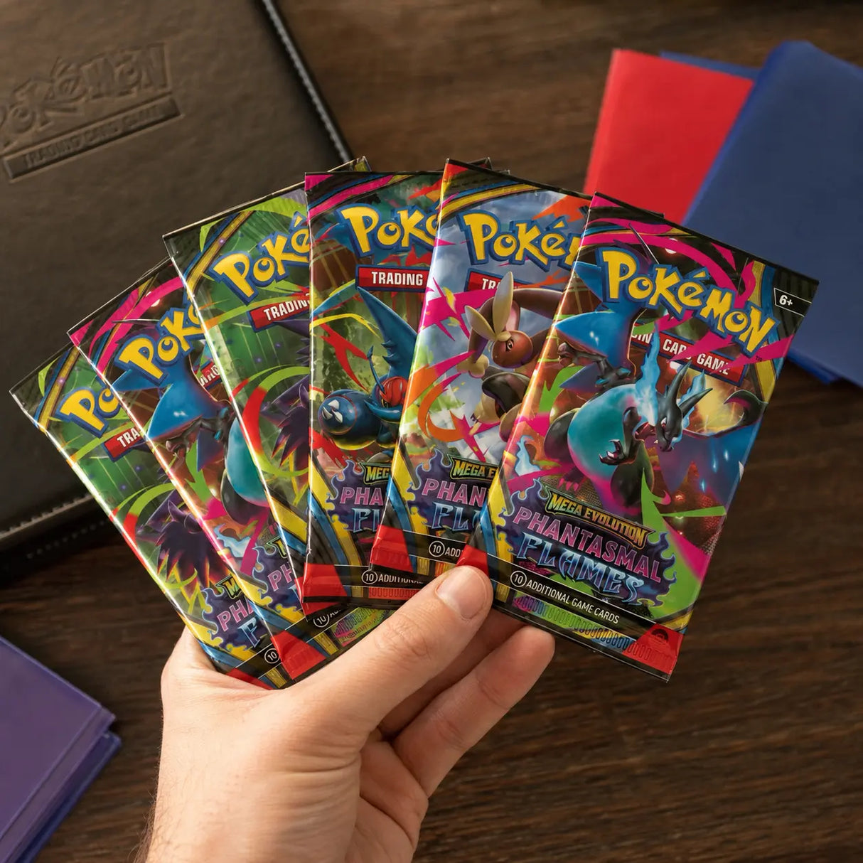 Hand hält 6 Phantasmal Flames Booster Packs aufgefächert vor Pokemon TCG Binder und Kartenhüllen