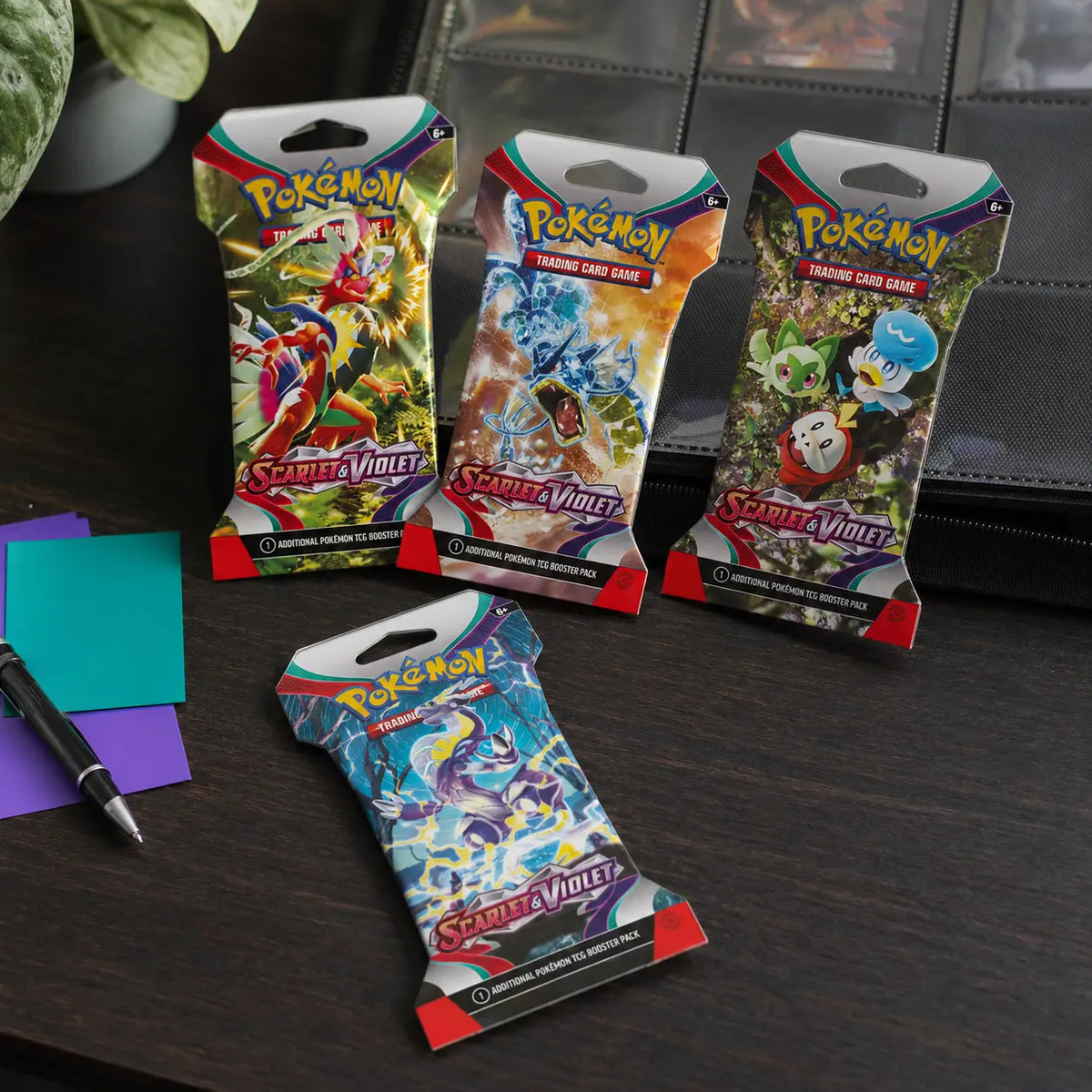 Vier Pokemon Scarlet & Violet Sleeved Booster Packs auf einem Schreibtisch mit Pokemon-Binder im Hintergrund